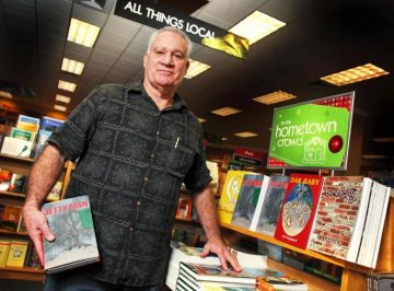 older gentleman with grey hair and black button upshirt stands next to stacks of books
